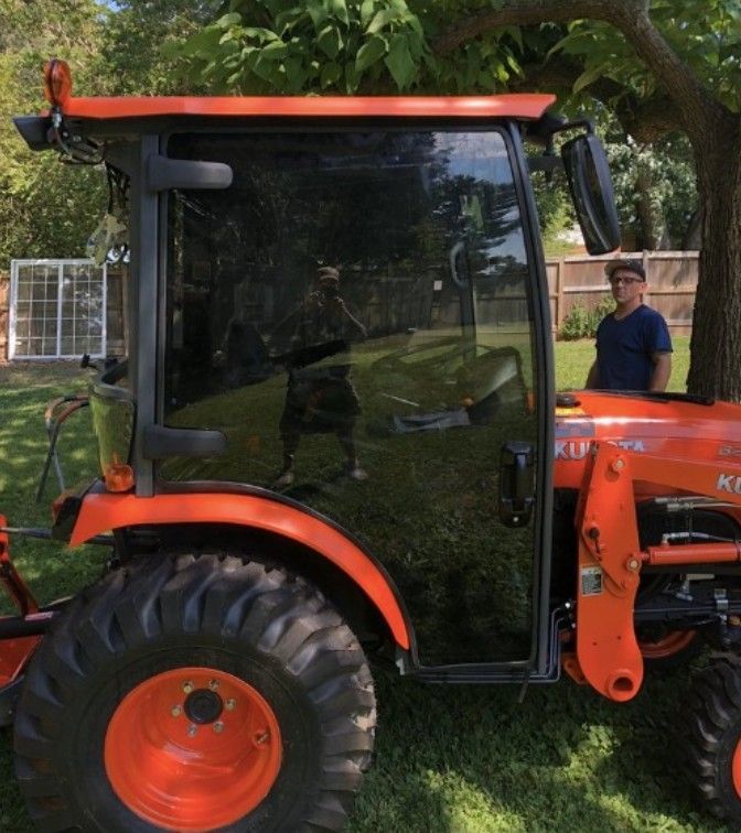 Orange Kubota tractor with a black enclosed cab. A person stands beside it in a backyard.