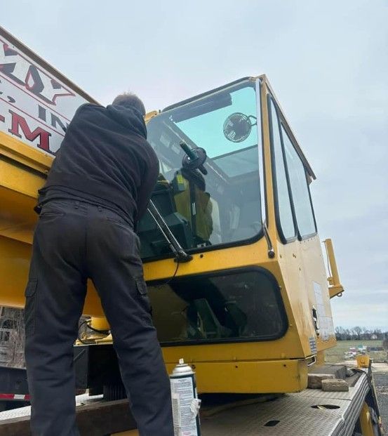 A person working on the window of a yellow crane cab outdoors.
