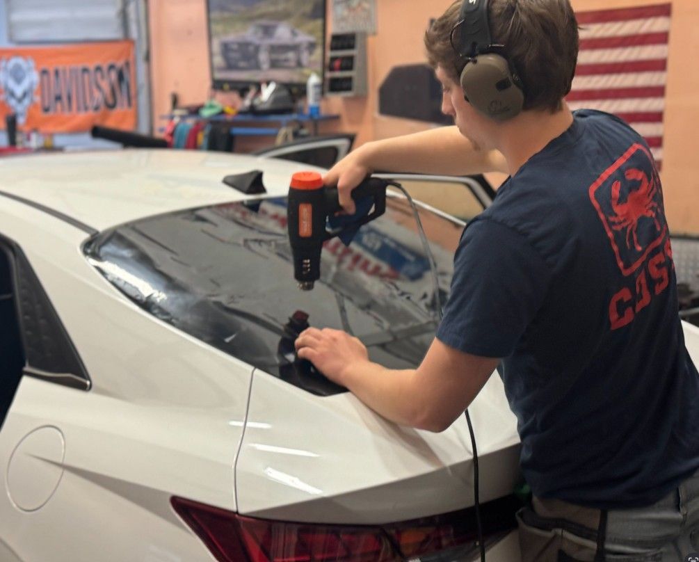 Man applying window tint to a white car with a heat gun in a shop. He is wearing ear protection.