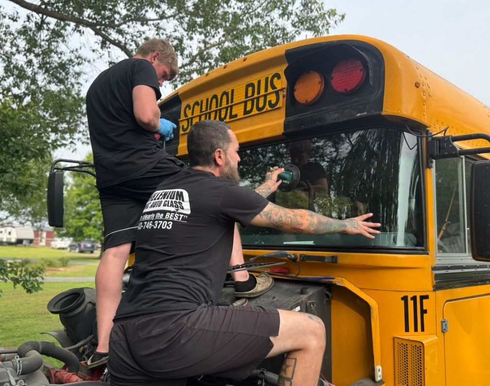 Two people replace a school bus windshield; yellow bus, cloudy sky, outdoor setting.
