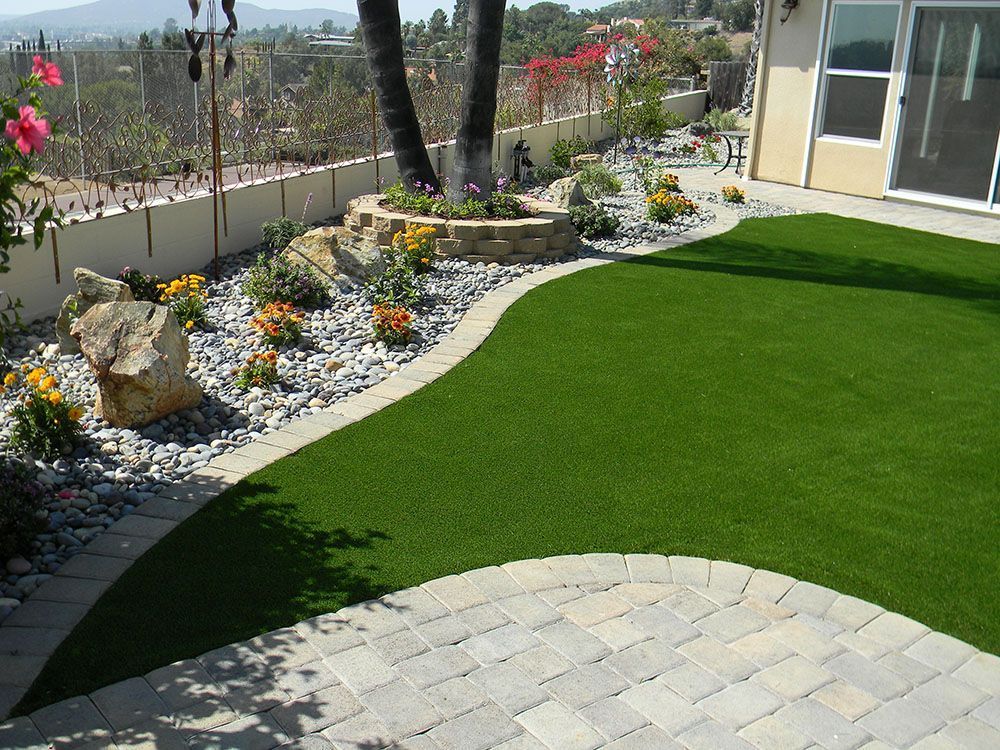 Landscaped backyard with bright green lawn, stone patio, flower beds, and a view of hills beyond the fence