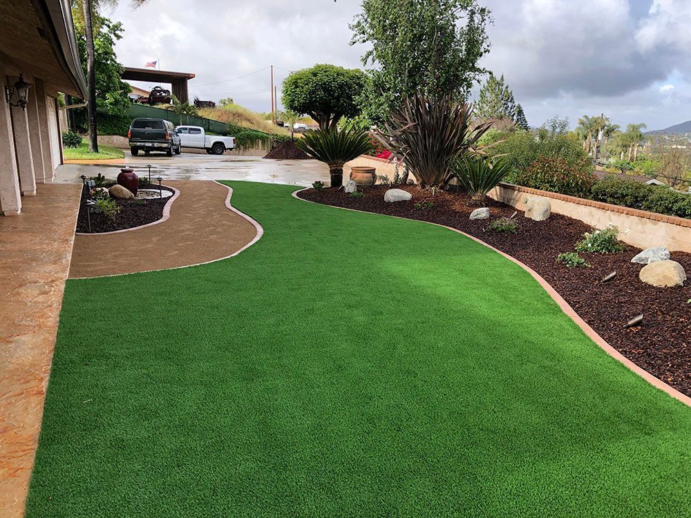 Landscaped front yard with artificial turf, curved flower beds, shrubs, and a parked car under cloudy skies