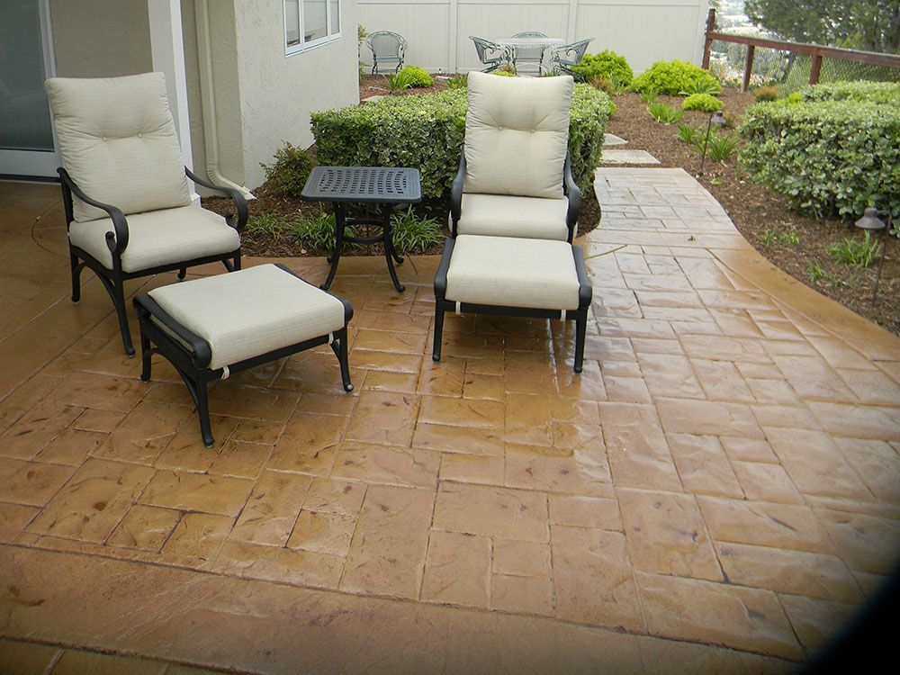 Patio seating area with two cushioned chairs, an ottoman, and a small table on a wet stone patio.