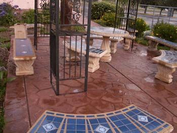 Outdoor patio with stone benches and tables on a wet brick floor, seen through a black metal gate