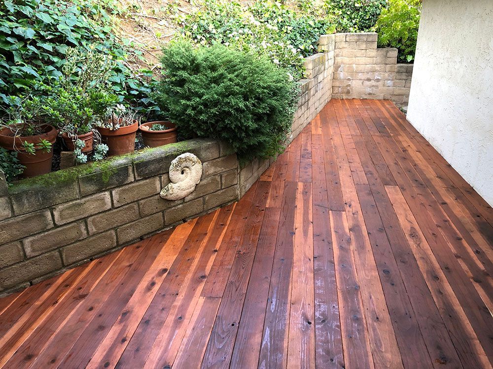 Wooden deck patio with potted plants and a stone retaining wall beside a white house