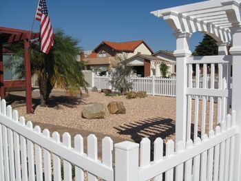 White picket fence around a desert yard with palm tree, flag, and white house in the background