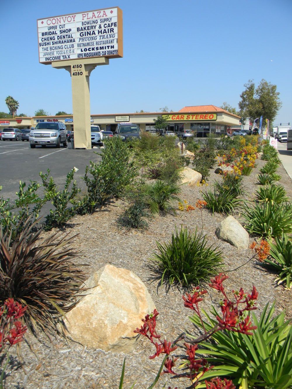 Desert-style shopping center with landscaped shrubs, rocks, and a tall roadside sign under a blue sky