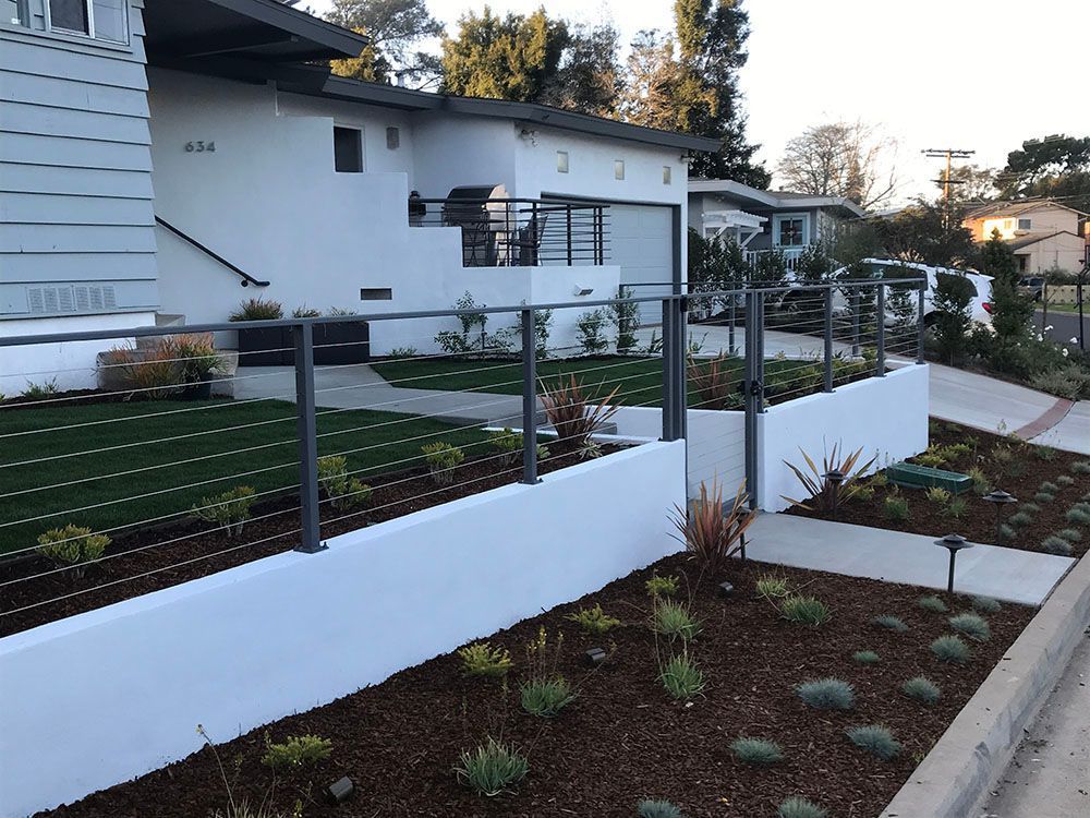 Modern white house with a landscaped front yard, low white wall, and metal fence along the sidewalk.
