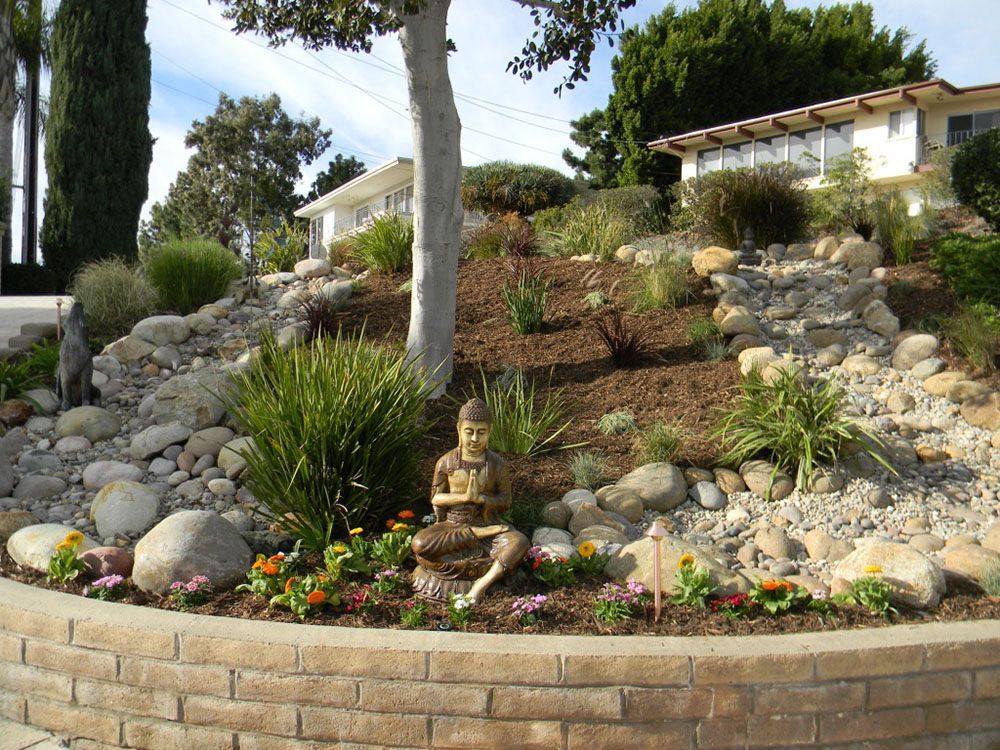 Tiered rock garden with succulents, flowers, a small Buddha statue, and houses in the background