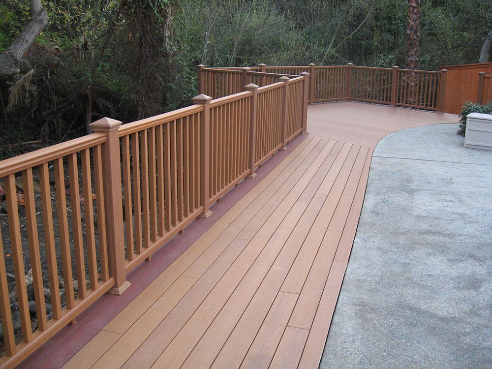 Curved wooden deck with railing beside a concrete patio, overlooking trees and greenery