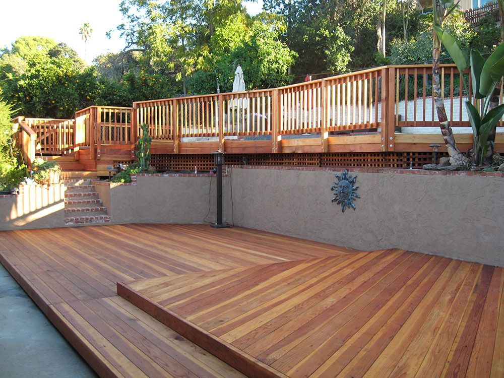 Wooden deck and stairs leading to a raised patio in a sunny backyard with trees and stone wall