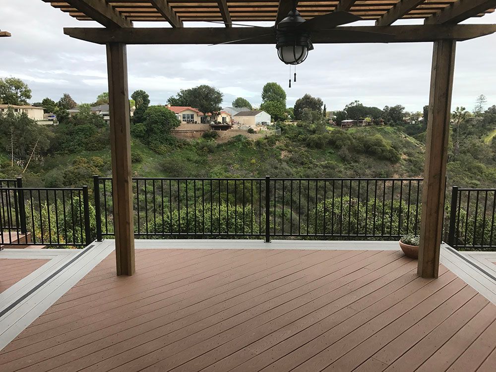 Covered wooden deck overlooking a green hillside and houses beyond a black metal railing.
