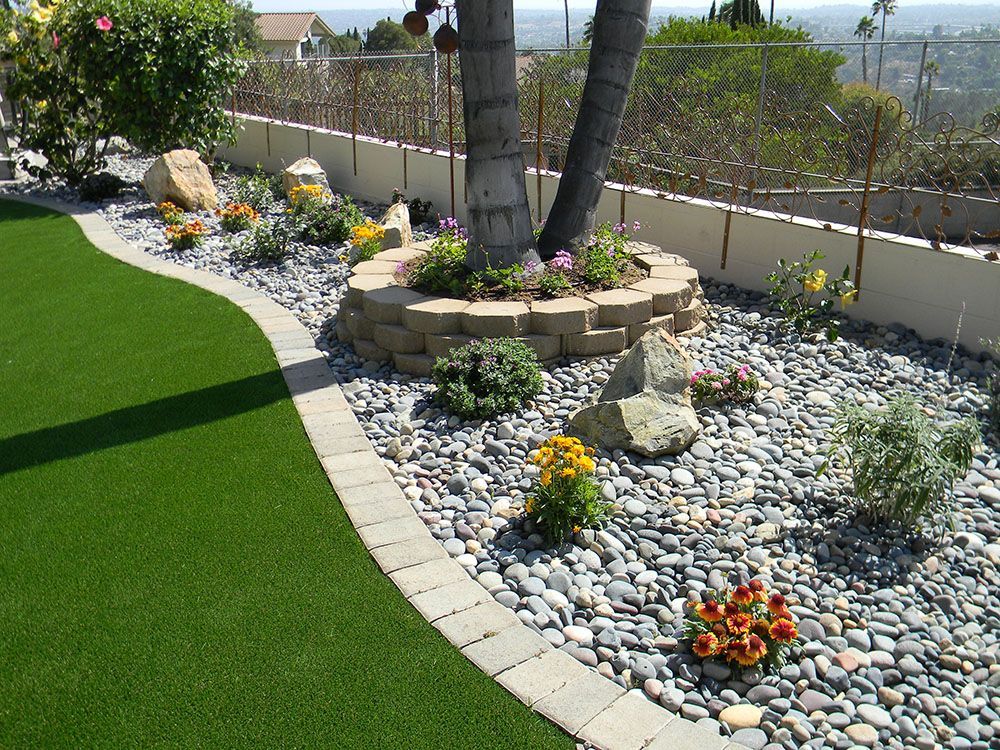 Curved landscaped garden with artificial grass, river rocks, flowers, and palm trees along a fence