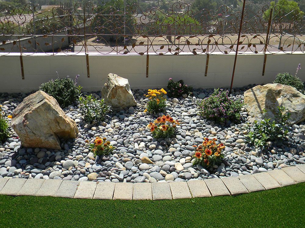 Rock garden with large stones, small flowering shrubs, and a curved border under bright sunlight