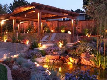 Illuminated landscaped garden with a wooden pergola, stone steps, and a reflecting pond at dusk