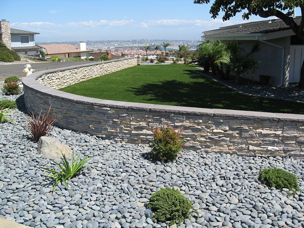 Curved stone retaining wall bordering a green lawn and rock garden with a distant city view