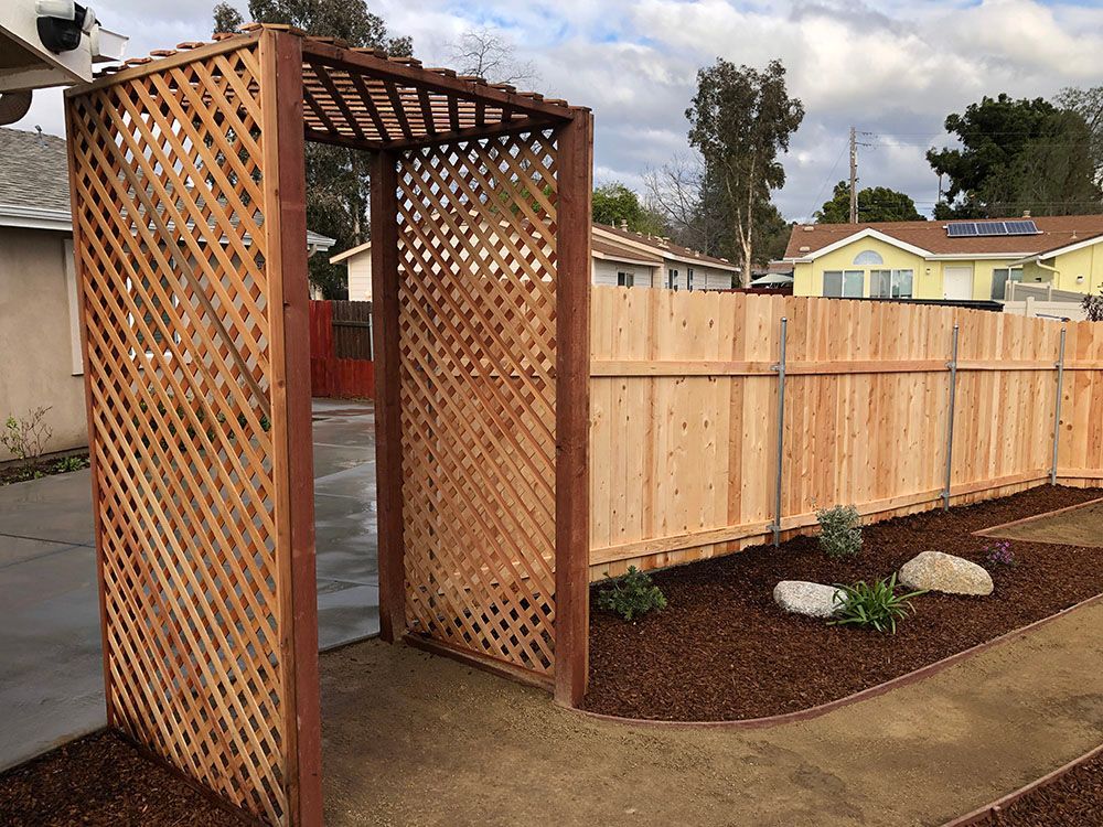 Wooden garden trellis entrance beside a fenced yard with mulch, rocks, and a small tree.