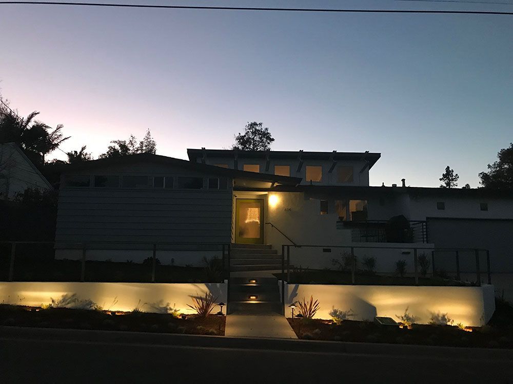 Modern house at dusk with warm porch lights and a lit walkway, silhouetted against a twilight sky
