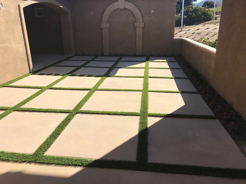 Sunlit patio with square stone pavers separated by green grass strips beside tan stucco walls