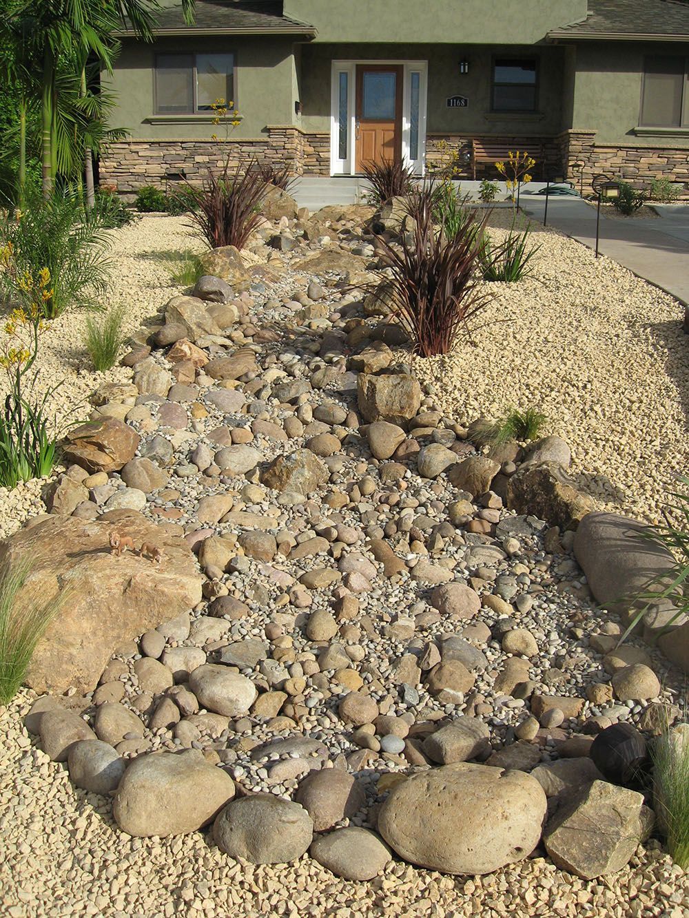 Rock garden with dry creek bed of stones leading to a house entrance and landscaped yard.