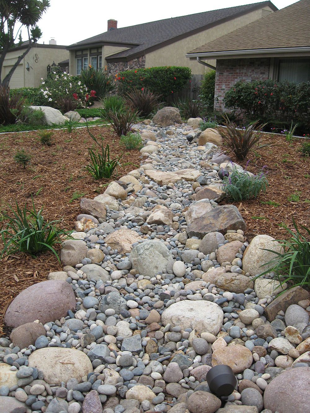 Rock-lined dry creek bed in a suburban front yard with shrubs and houses in the background