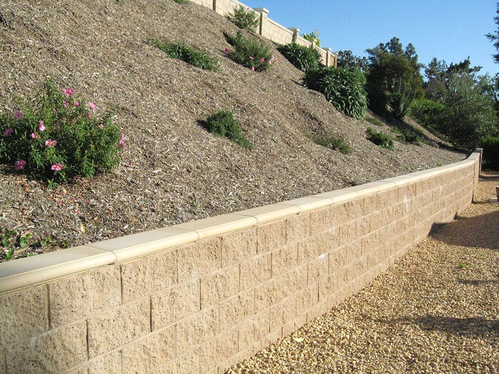 Retaining wall beside a sloped gravel hillside with small shrubs and trees under a blue sky