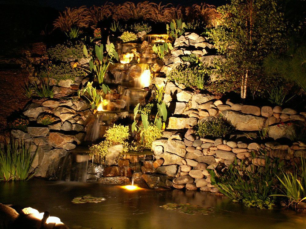 Lit garden waterfall cascading over rocks into a pond at night, illuminated by warm lights