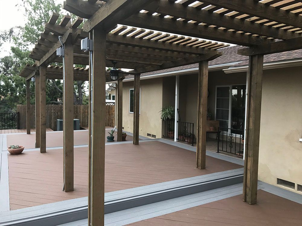 Wooden pergola over a tiled patio beside a beige house with sliding glass doors.