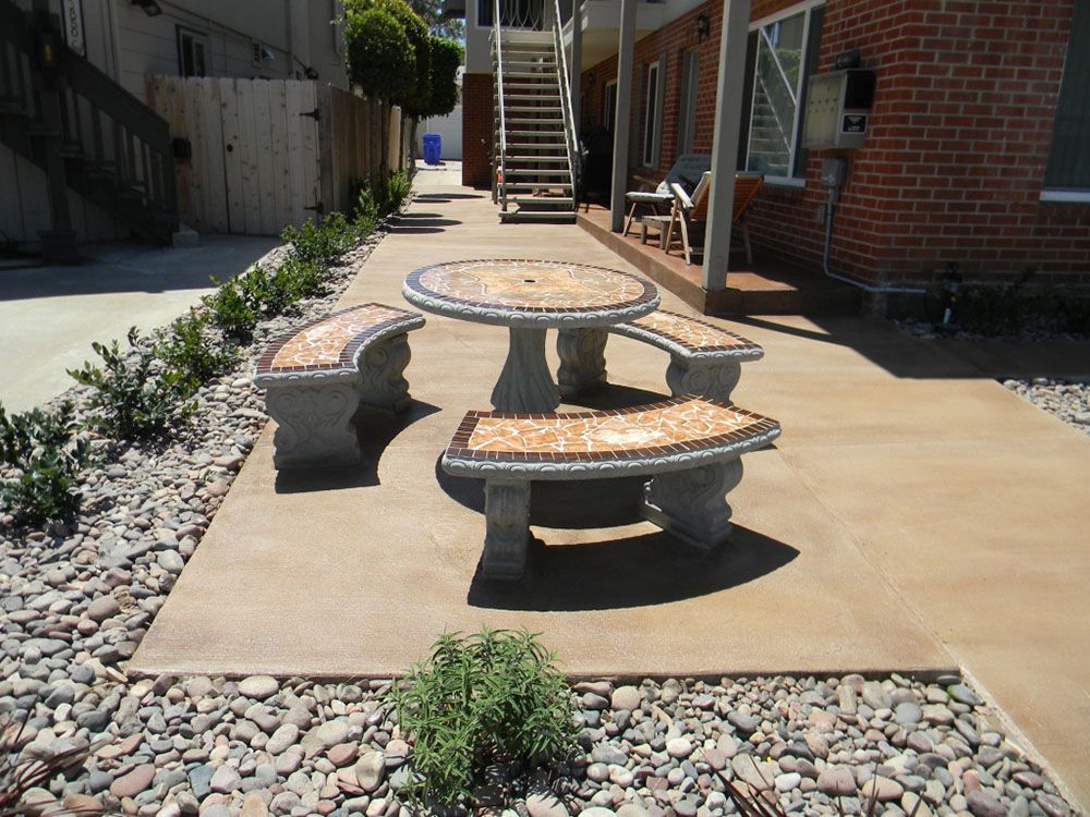 Outdoor picnic table and benches on a concrete patio beside a brick building and rock landscaping.
