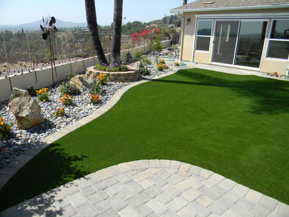 Backyard patio with artificial grass, stone pavers, and landscaped garden beside a house.