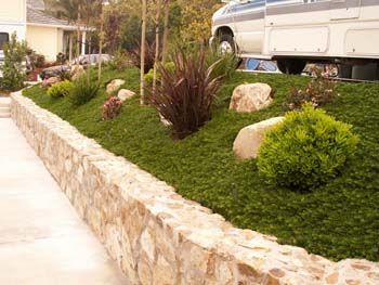 Stone retaining wall edging a landscaped garden with green shrubs and rocks beside a sidewalk and parked RV