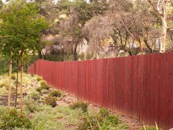 Red wooden fence beside a grassy path with trees and shrubs in a park-like area.