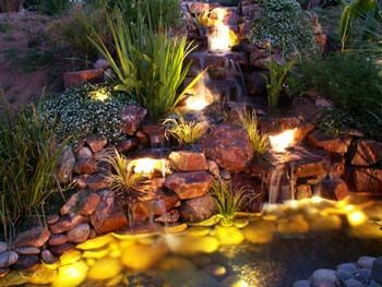 Rock waterfall cascading into a lit backyard pond at dusk, surrounded by plants and stones