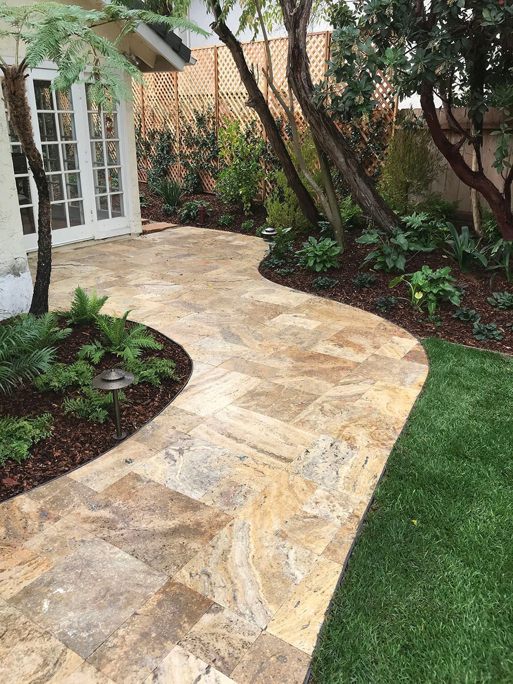 Stone garden path curving past a house entrance, bordered by green lawn and landscaped plants.