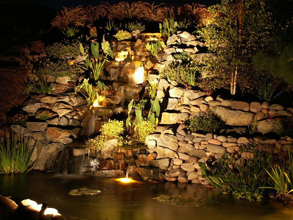 Lit rock waterfall cascading into a pond at night, surrounded by greenery and warm lights.