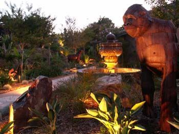 Illuminated garden with a large dinosaur statue beside a lit fountain at dusk