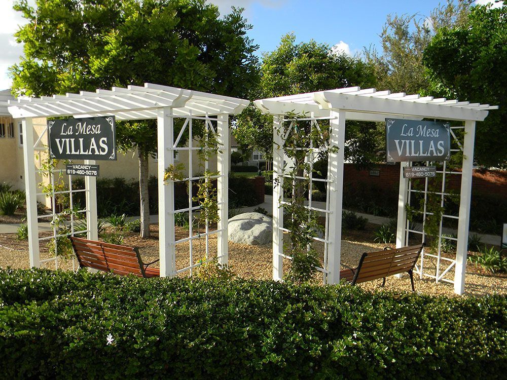 Two white pergolas with benches and “Le Mont VILLAS” signs behind a hedge in a sunny garden