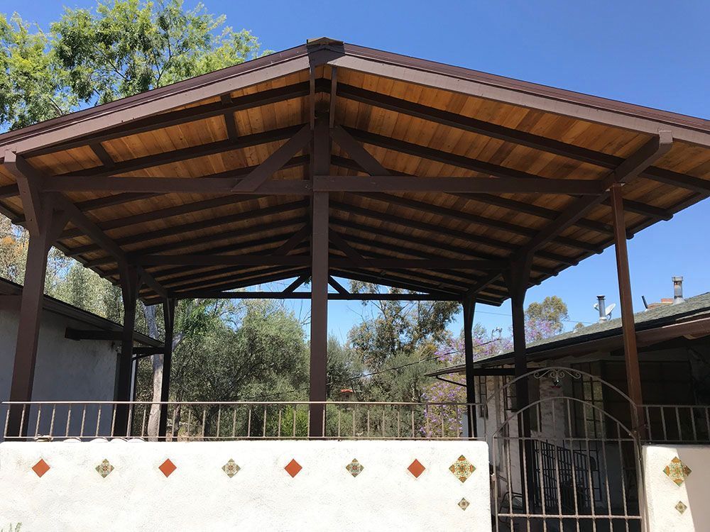 Covered wooden gazebo with open sides and a white patterned wall, set in a sunny outdoor yard