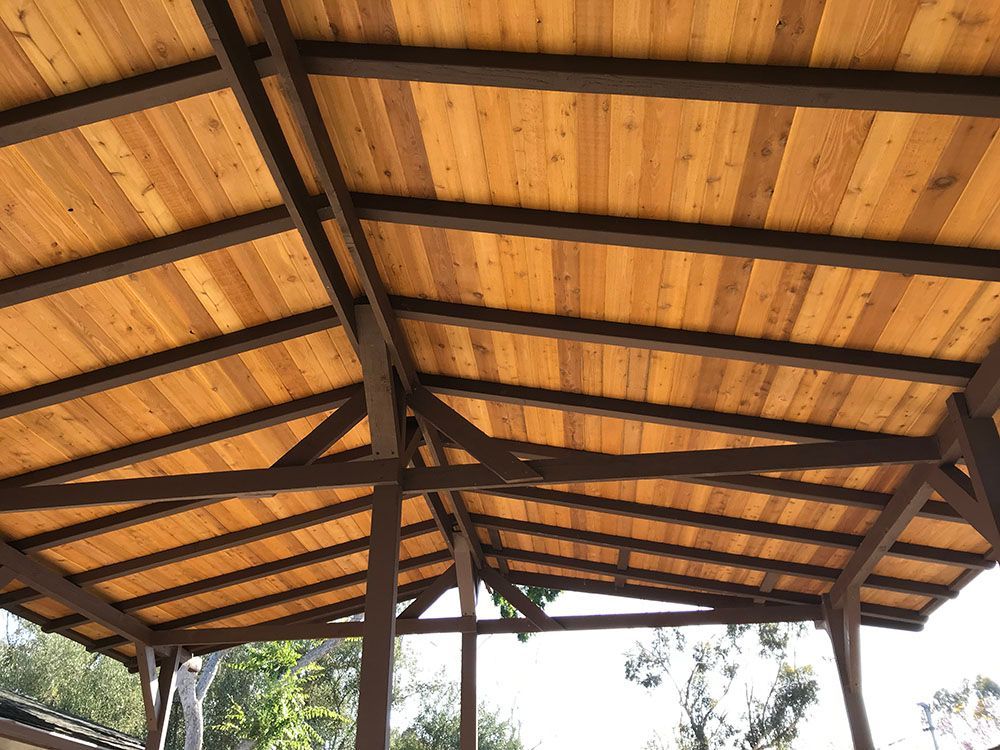 Wooden pavilion roof with dark beams and tan panels, viewed from below against a bright sky.