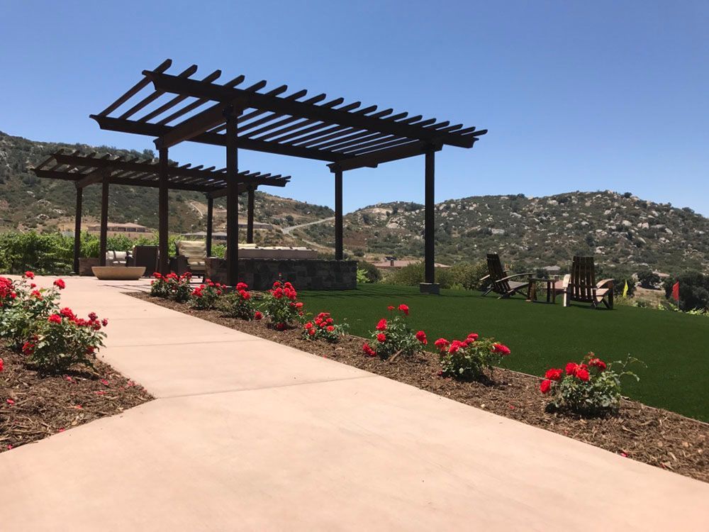 Garden patio with a pergola, red flowers along a path, and hills in the background.
