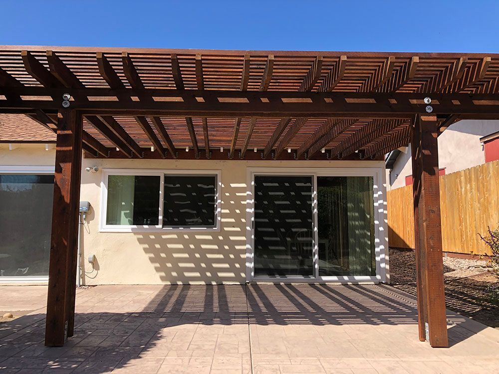Patio with wooden pergola casting striped shadows over a beige house with sliding glass doors.