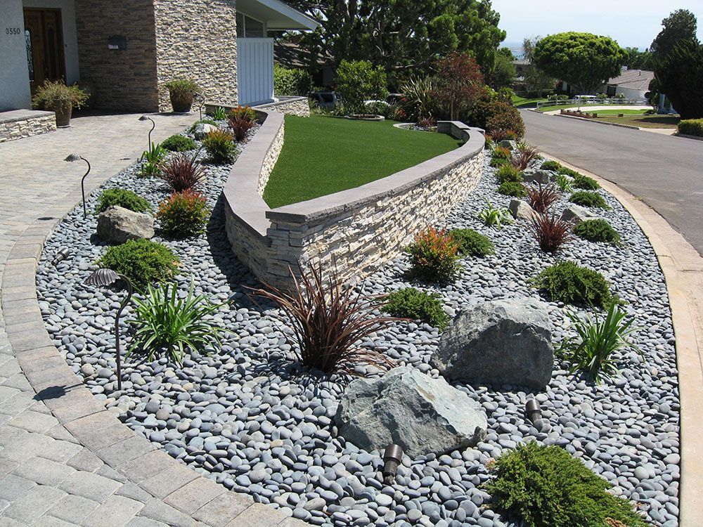 Landscaped front yard with stone retaining wall, white rocks, shrubs, and green lawn beside a street.