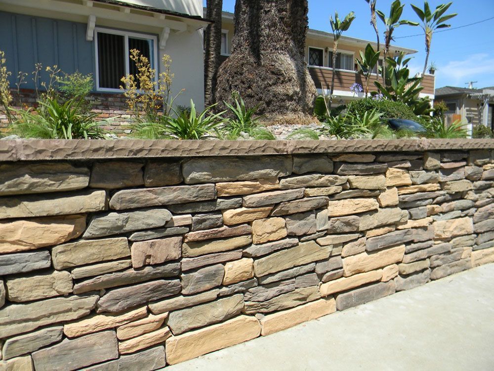 Stone retaining wall beside a sidewalk with a palm tree and houses in the background
