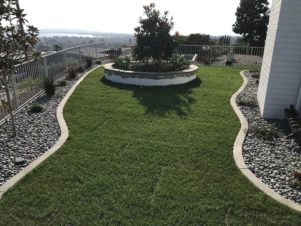 Curved green lawn bordered by white rocks, with a circular planter and distant hillside view.