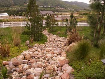 Rock garden path leading toward a white fence, with trees, grasses, and hills in the background.
