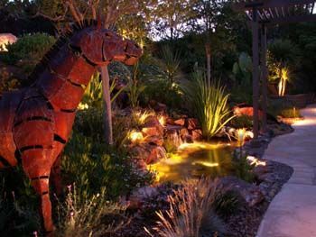 Sculpted animal silhouette in a lit garden with tropical plants and a curving path at dusk