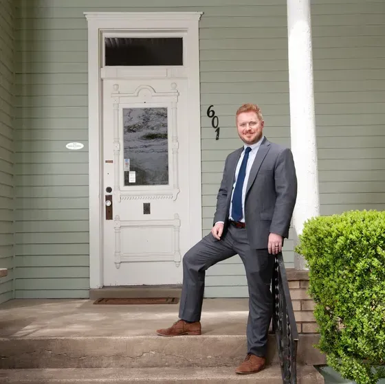 Man in gray suit stands on porch steps in front of a light green house.