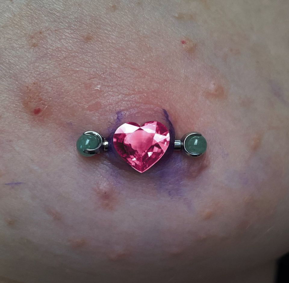 A close-up of a belly button piercing with a pink heart-shaped stone.