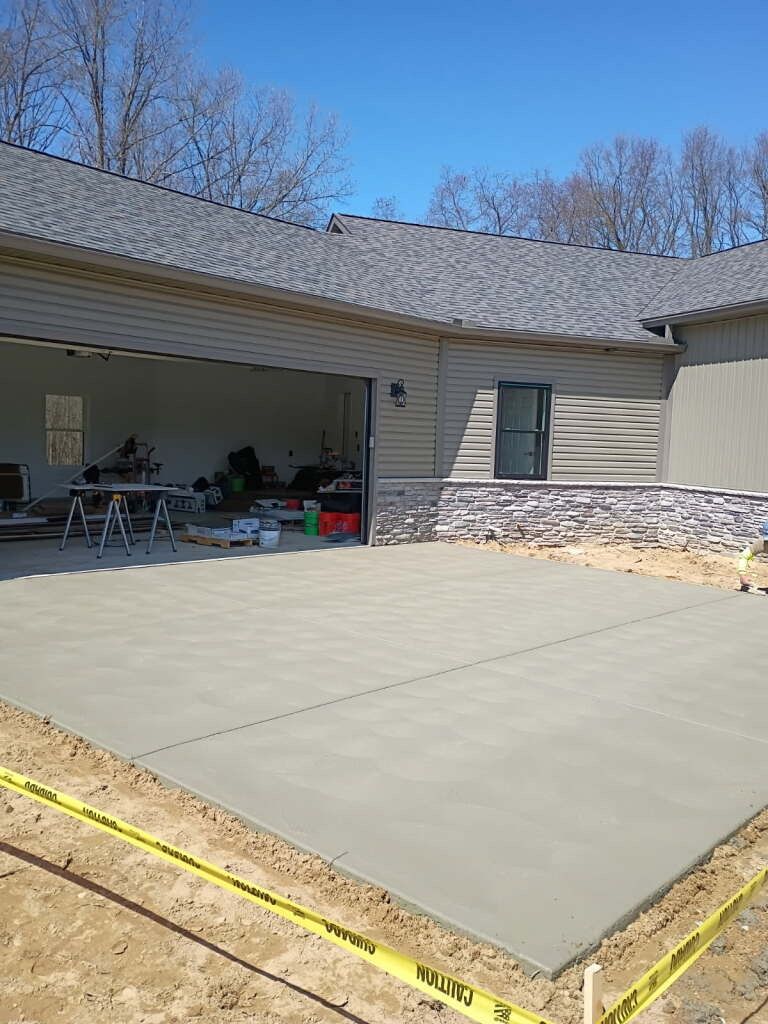 New concrete patio beside a beige house with an open garage and stone foundation.