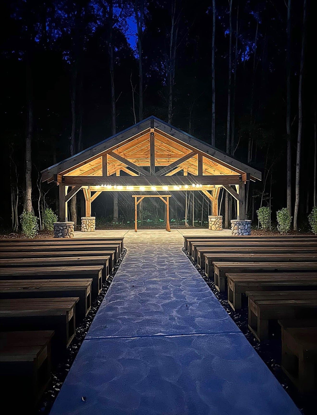 Night-lit outdoor chapel with wooden pews and a central aisle leading to a lit pavilion